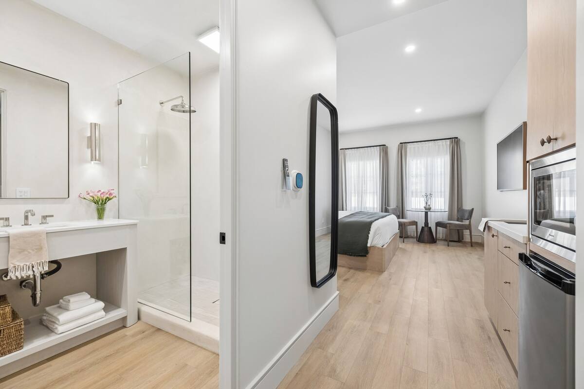 Moderni hotellihuone, jossa on suihku, suuri peili, ruokailutila pöytineen ja tuoleineen sekä keittiö, jossa on jääkaappi.