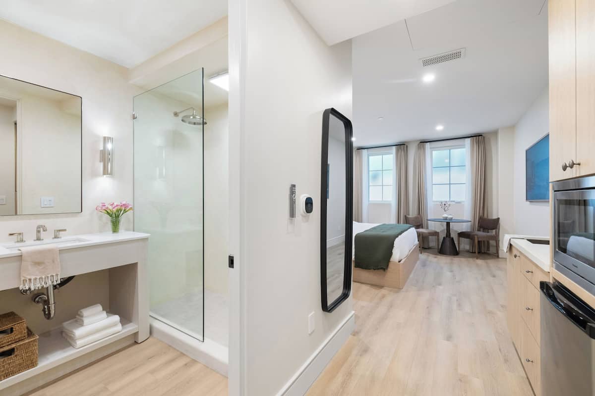 Moderni hotellihuone, jossa on suihku, suuri peili, sänky, ruokailutila ja keittiö.