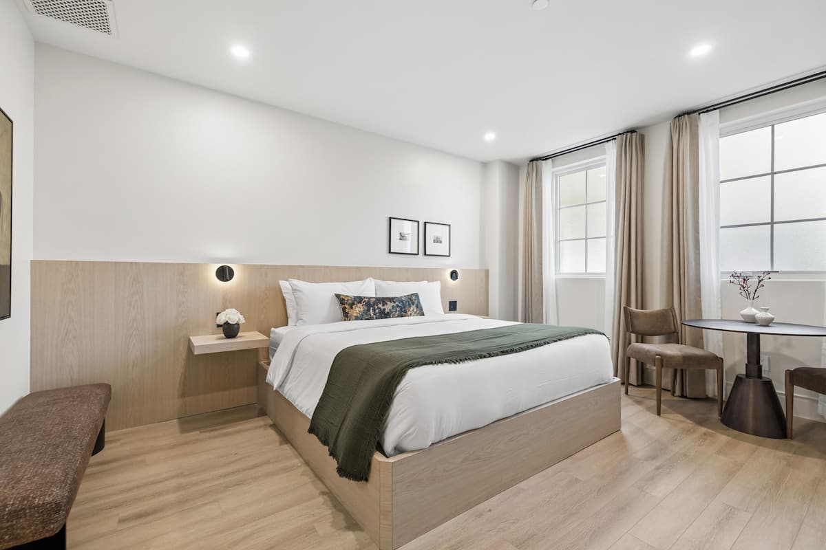 Moderni hotellihuone, jossa on suuri sänky, yöpöydät, penkki, pieni pöytä tuoleineen ja ikkuna, jossa on verhot.