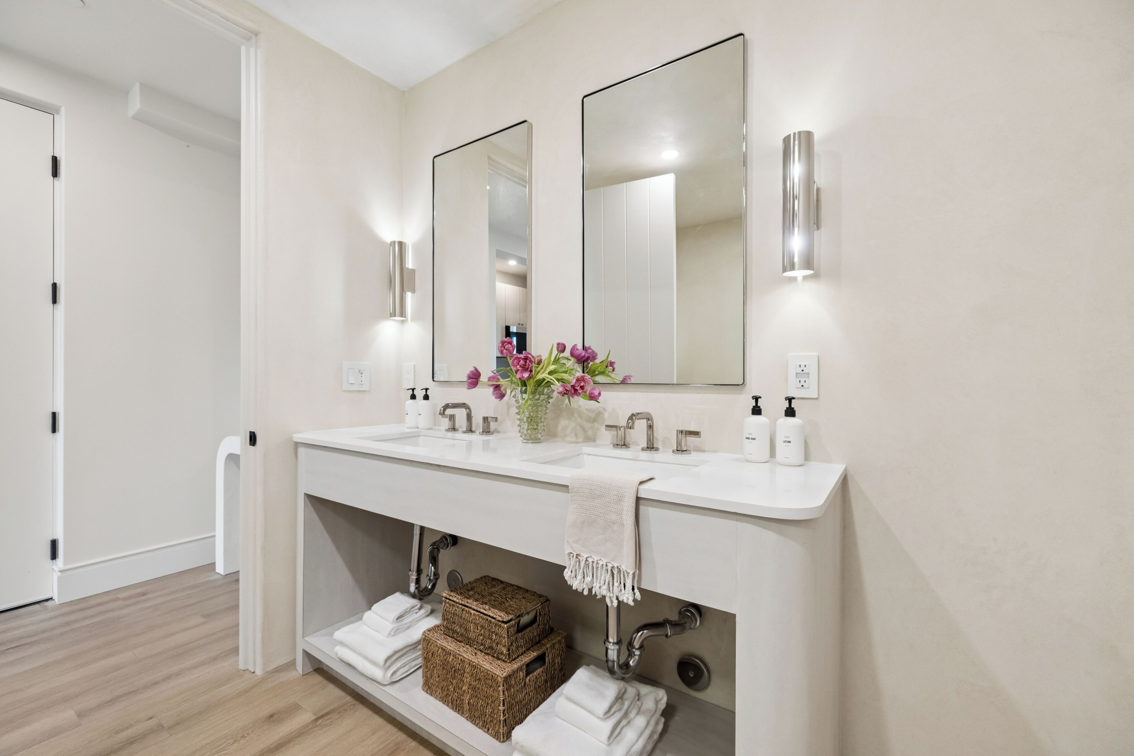 A modern bathroom with a white vanity, two mirrors, a vase with flowers, and neatly folded towels.