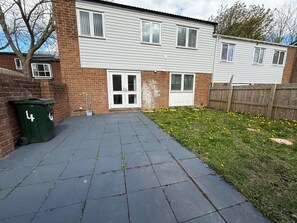 A residential building with a paved driveway, a green trash bin, and a grassy area with yellow flowers. - “Large Newcastle Home for 8– 4 guest Bedrooms, 5 Beds (Newcastle upon Tyne)