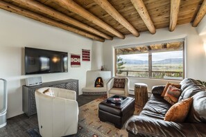 A living room with a fireplace, a large window with a view, a flat-screen TV, and a wooden ceiling.
