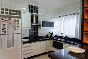A modern kitchen with white cabinets, a black countertop, a stainless steel range hood, and a window with white curtains. - Casa c Churrasqueira a 300m da Praia de Massagua u (Caraguatatuba)