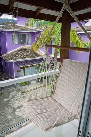 A hammock chair suspended from a ceiling, a purple building in the background, and a tiled patio area. - Casa c Churrasqueira a 300m da Praia de Massagua u (Caraguatatuba)