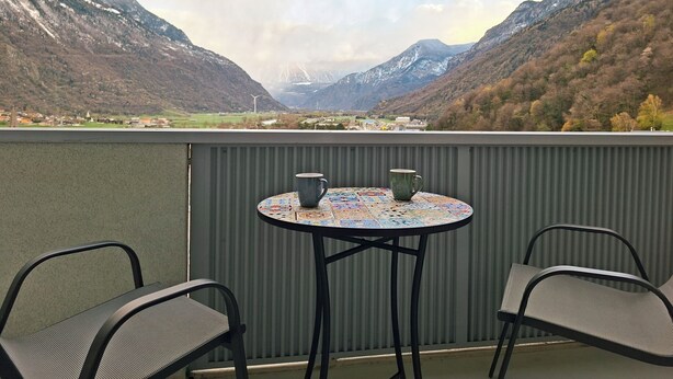 Een balkon met een tafel en stoelen, uitkijkend op een bergachtig landschap met een dorp in de verte.
