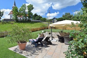 Eine Terrasse mit Gartenmöbeln, einem Blumentopf und Blick auf eine Kirche sowie umliegendes Grün.