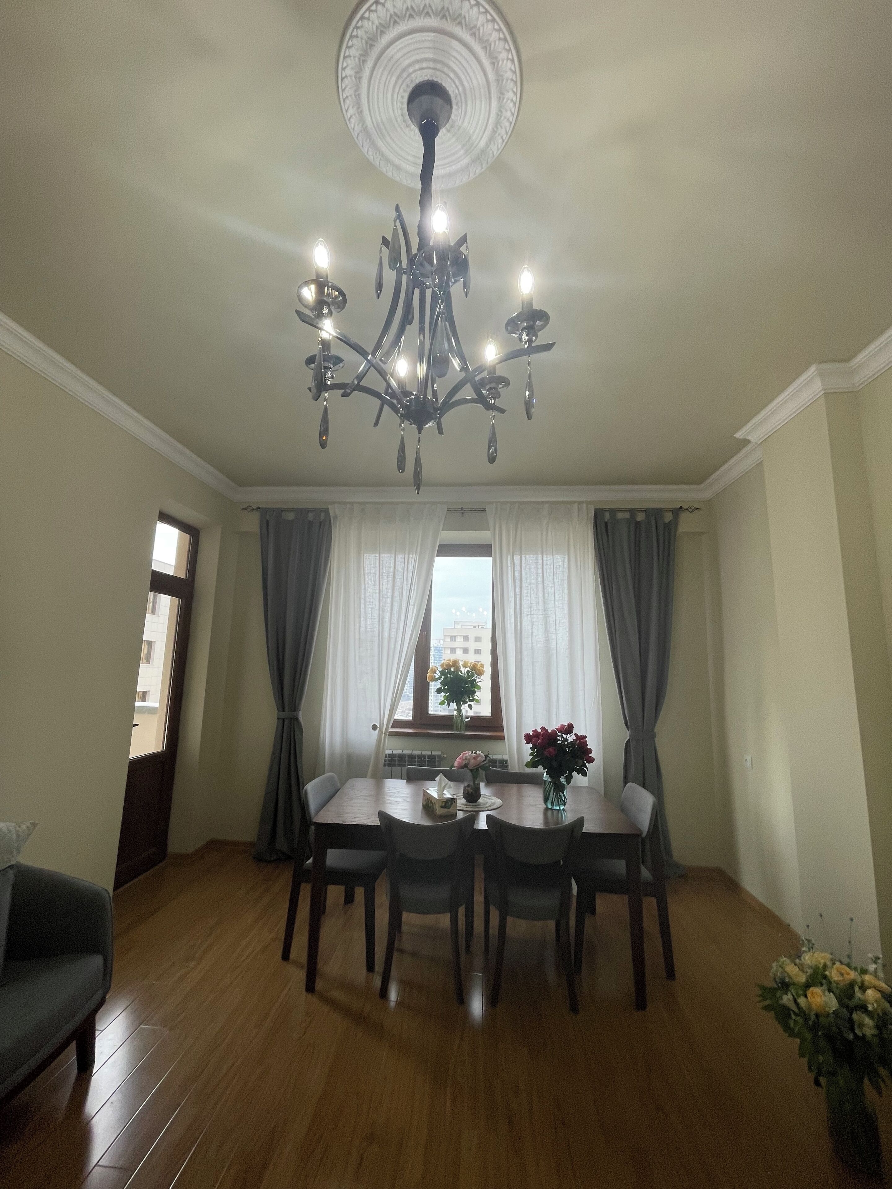 Une salle à manger avec un lustre, une table entourée de chaises, un vase rempli de fleurs et une fenêtre ornée de rideaux.