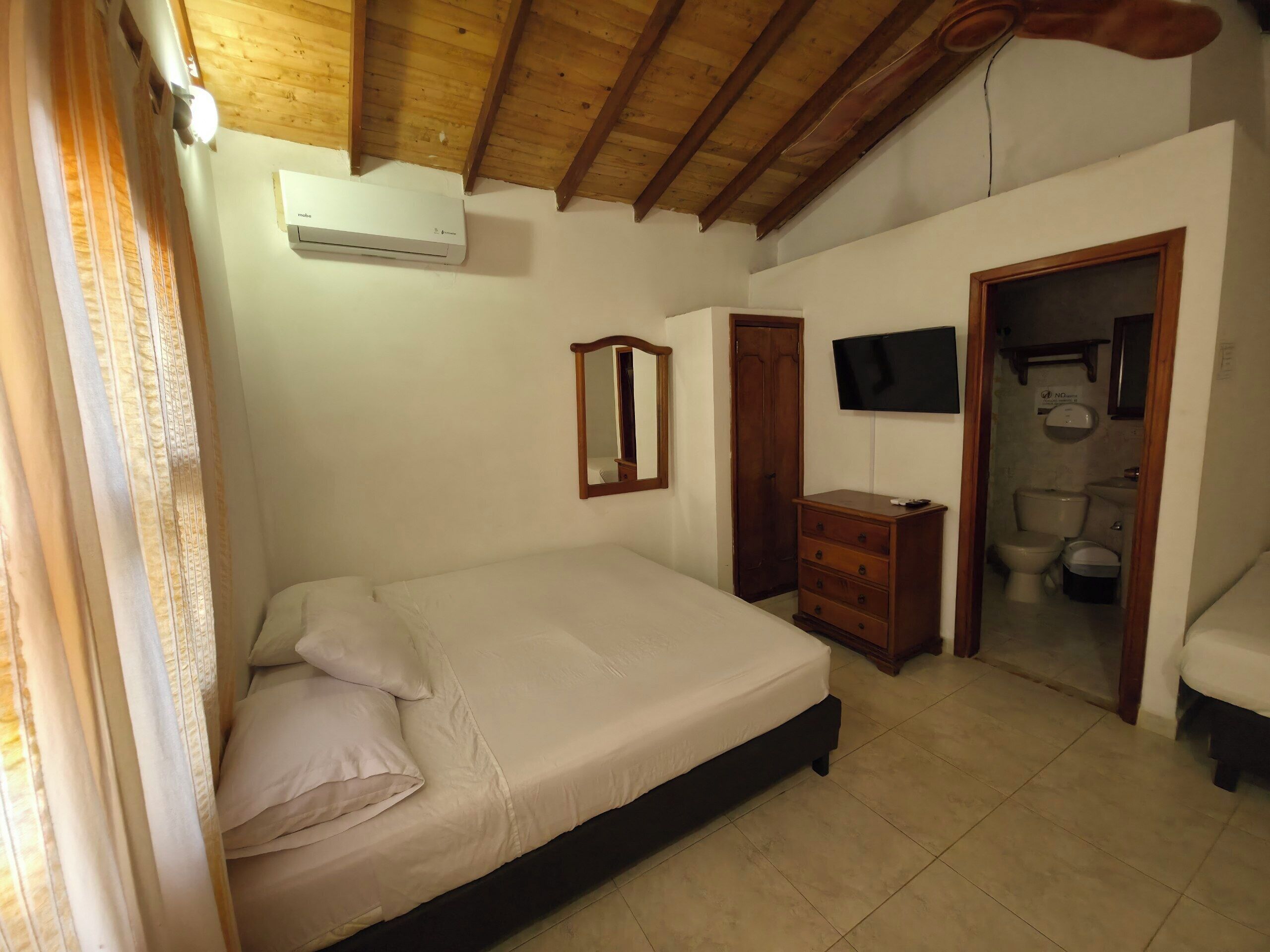 Een slaapkamer met een bed, een dressoir, een spiegel, een televisie en een badkamer, zichtbaar door een open deur.