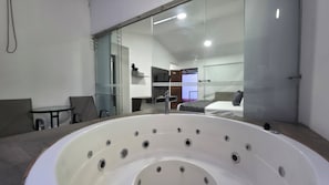 A modern bathroom with a large bathtub, a glass shower enclosure, and a visible bedroom with a bed and a TV.