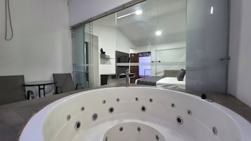 Un baño moderno con una bañera grande, un box de ducha de vidrio y se ve un dormitorio con cama y televisor.