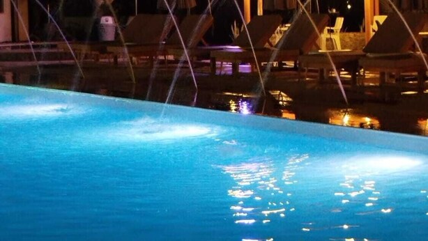Un espace piscine nocturne doté de jets d’eau, de chaises longues et d’un espace salon abrité.