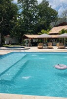 Una piscina con sillones de descanso y sombrillas, rodeada de árboles y un edificio con techo de tejas.