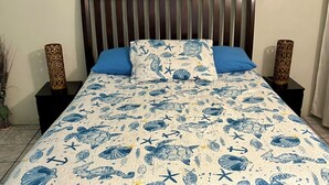 Un llit amb un llençol blau i blanc de temàtica marinera, un panell frontal de fusta, dues taules de nit amb vasses decoratives i una làmpada fixada a la paret.