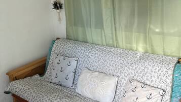 Una habitación con un sofá cama de madera, una cortina verde, un adorno de macramé en la pared y un cuadro con temática de playa.