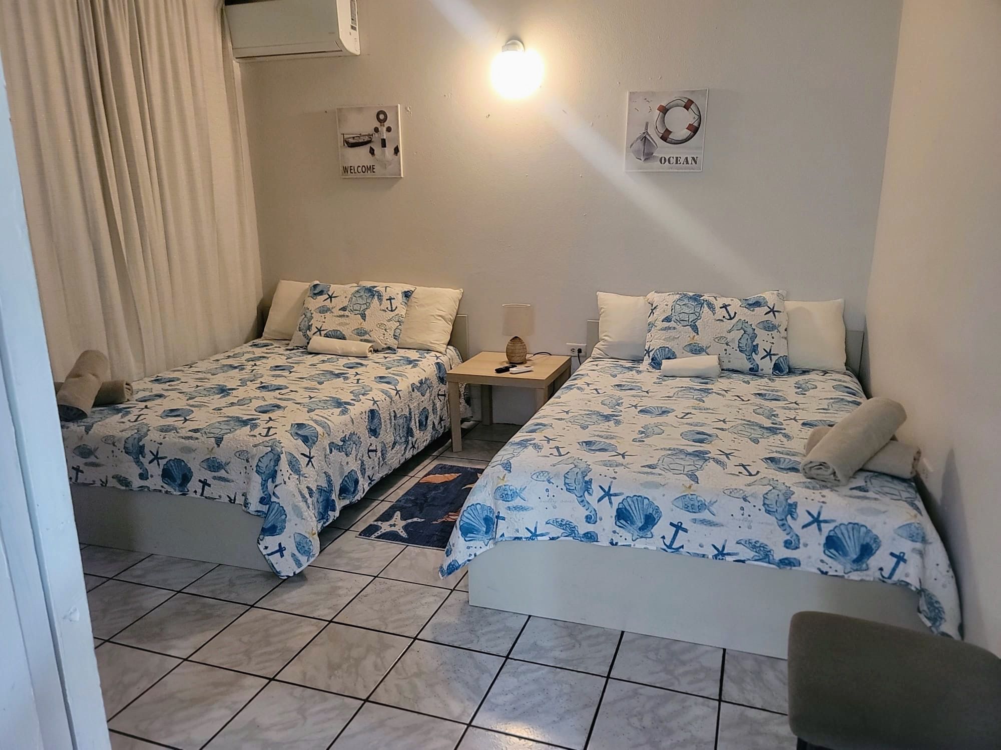 Ein Zimmer mit zwei Betten, jedes mit einem Bezug im Meeresstil, einem kleinen Tisch mit Lampe und einem Wandschild mit der Aufschrift 'WELCOME'.