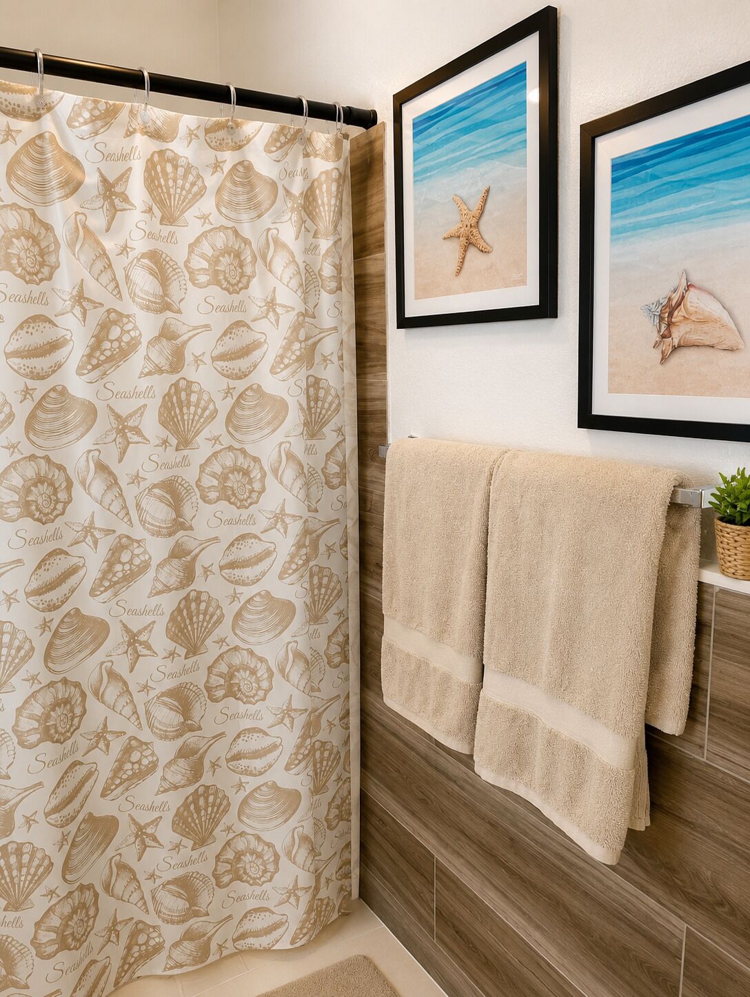A bathroom with a shower curtain featuring seashells and starfish, two framed pictures on the wall, and a towel rack with two towels.