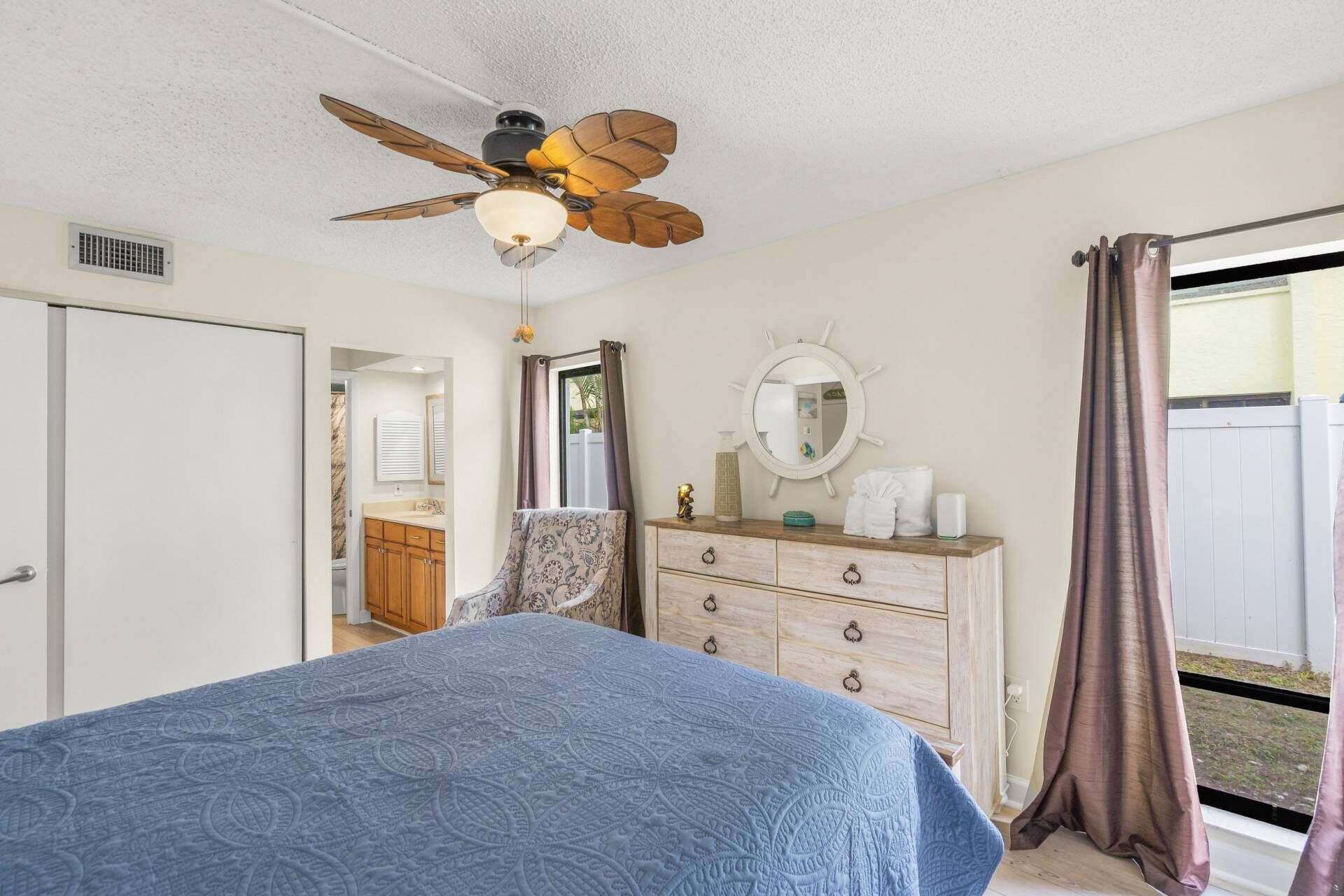 A bedroom with a bed, dresser, mirror, ceiling fan, and a view of the outdoors.
