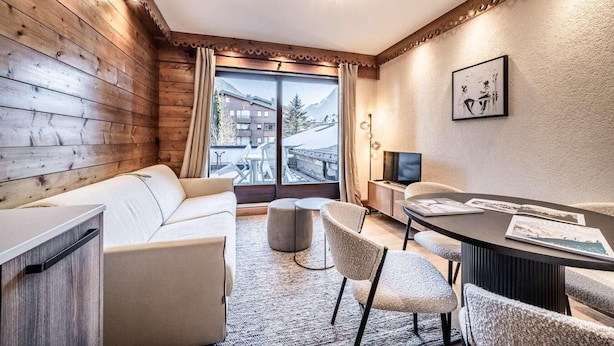 Quarto moderno com parede de madeira, sofá, mesa de jantar com cadeiras, TV e vista para uma montanha.