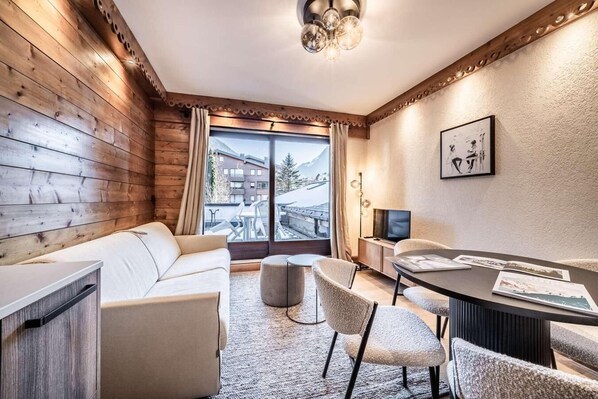 Ein modernes Zimmer mit einer Holzwand, einer Couch, einem Esstisch mit Stühlen, einem Fernseher und Blick auf einen Berg.