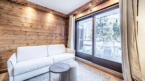 Quarto com parede de madeira, sofá branco, mesa redonda pequena e grande janela com vista para área externa com mesas e cadeiras.