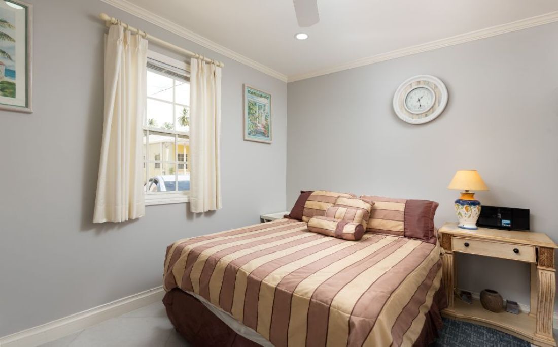 Een slaapkamer met een bed, nachtkastje met lamp, klok aan de muur en raam met gordijnen.