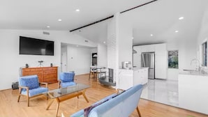 A modern living room with a glass coffee table, blue sofas, a wooden cabinet, and a flat-screen TV mounted on the wall. - 3BD 2BA House Breathtaking Views in Los Feliz Hills (Los Angeles)