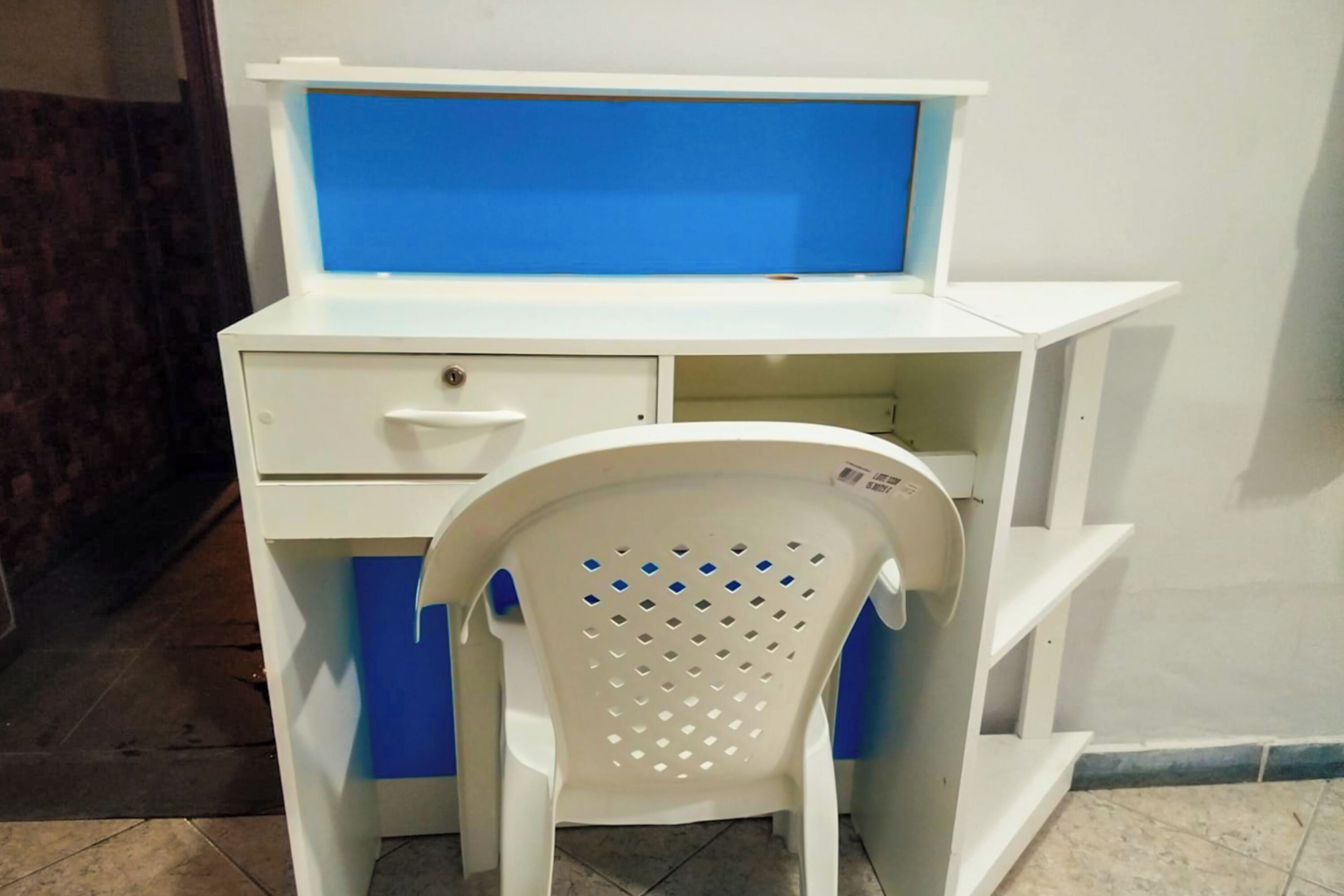 Un escritorio blanco con pantalla azul, una silla blanca con detalles azules y un piso de baldosas.
