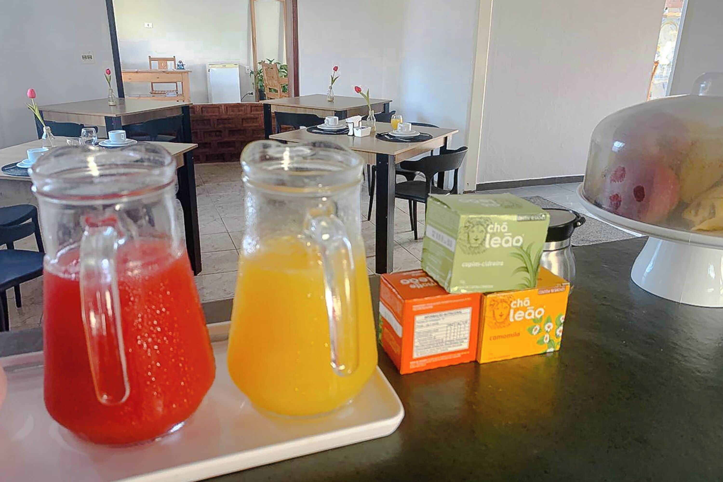 Disposição para o café da manhã com dois jarros de suco, uma cesta de frutas coberta e caixas de chá Leão.