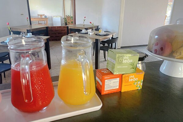 Disposição para o café da manhã com dois jarros de suco, uma cesta de frutas coberta e caixas de chá Leão.
