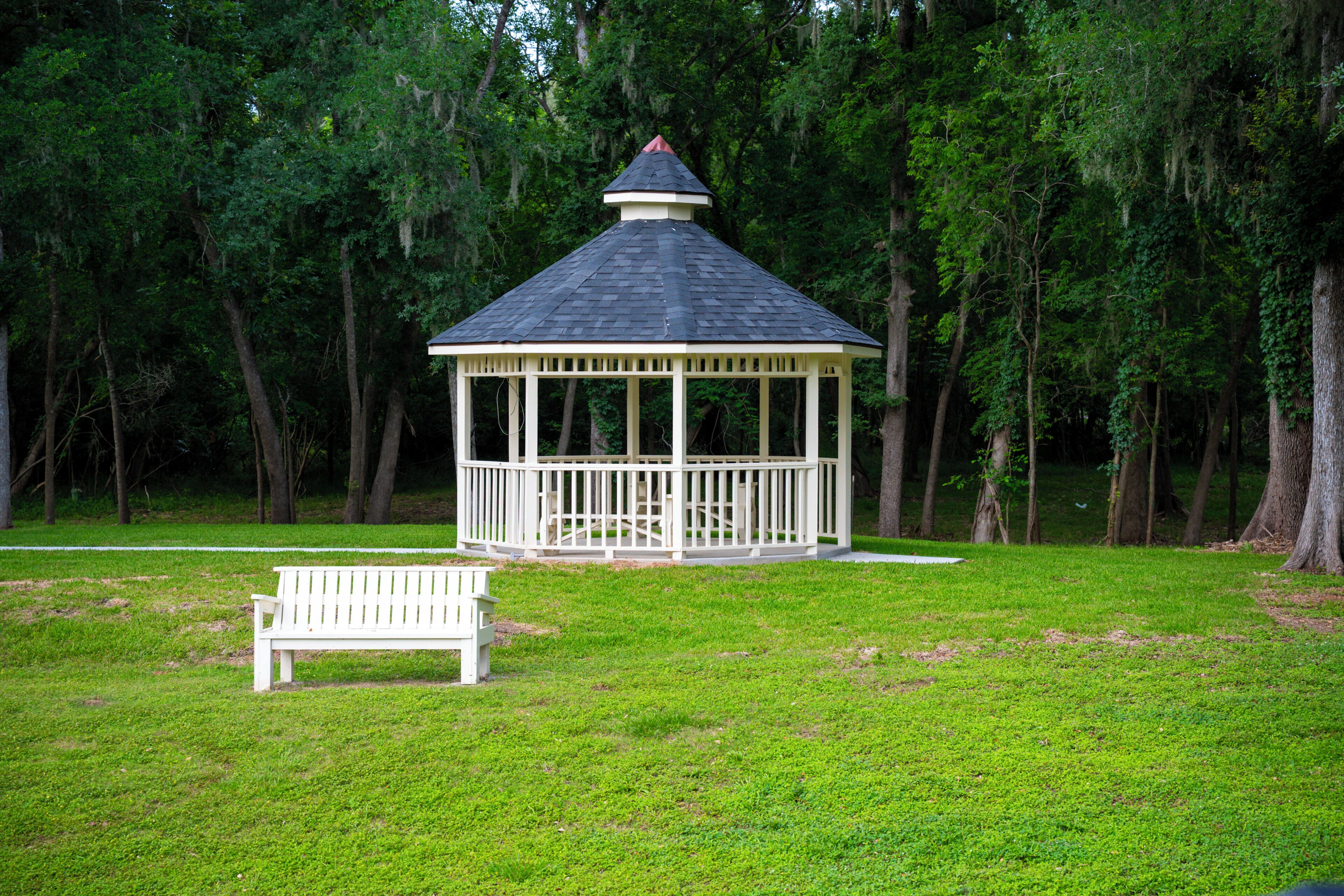 A gazebo with a white railing and a bench in front, surrounded by trees.