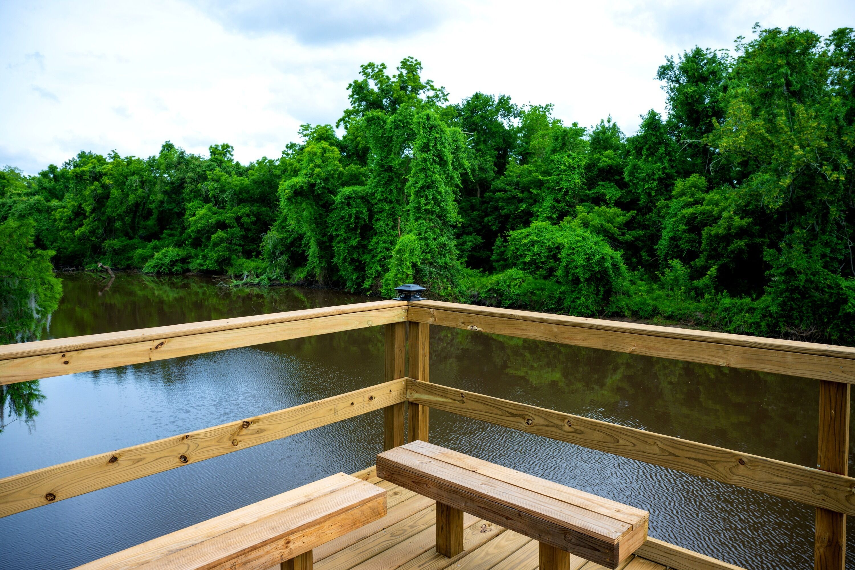 A wooden deck with a bench, overlooking a calm body of water and dense green foliage.