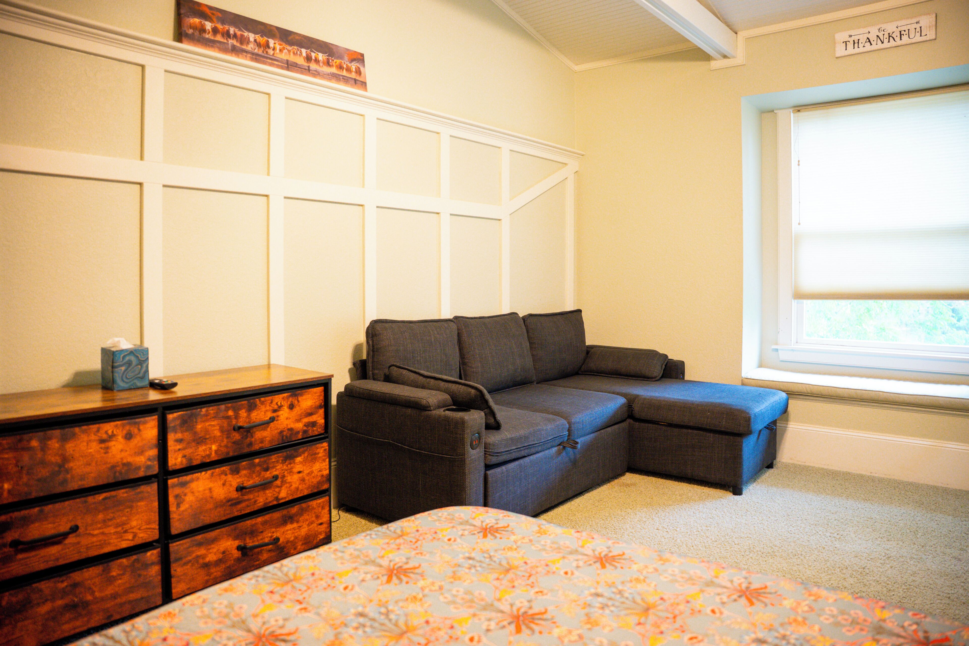 Un dormitorio con sofá seccional, cómoda de madera, cama con ropa de cama floral y ventana con vistas.