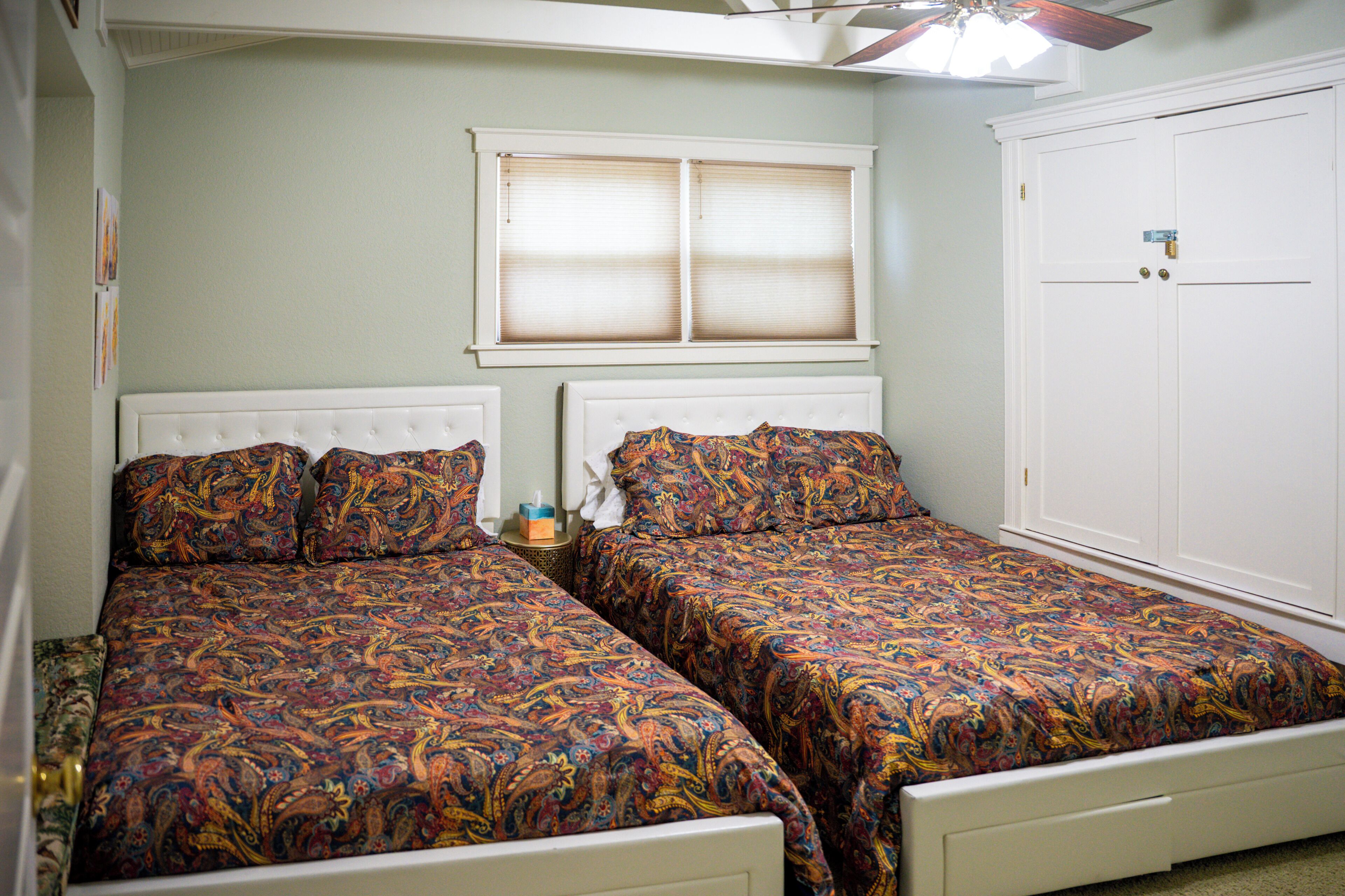 A bedroom with two beds, a window with blinds, a ceiling fan, and a wardrobe.