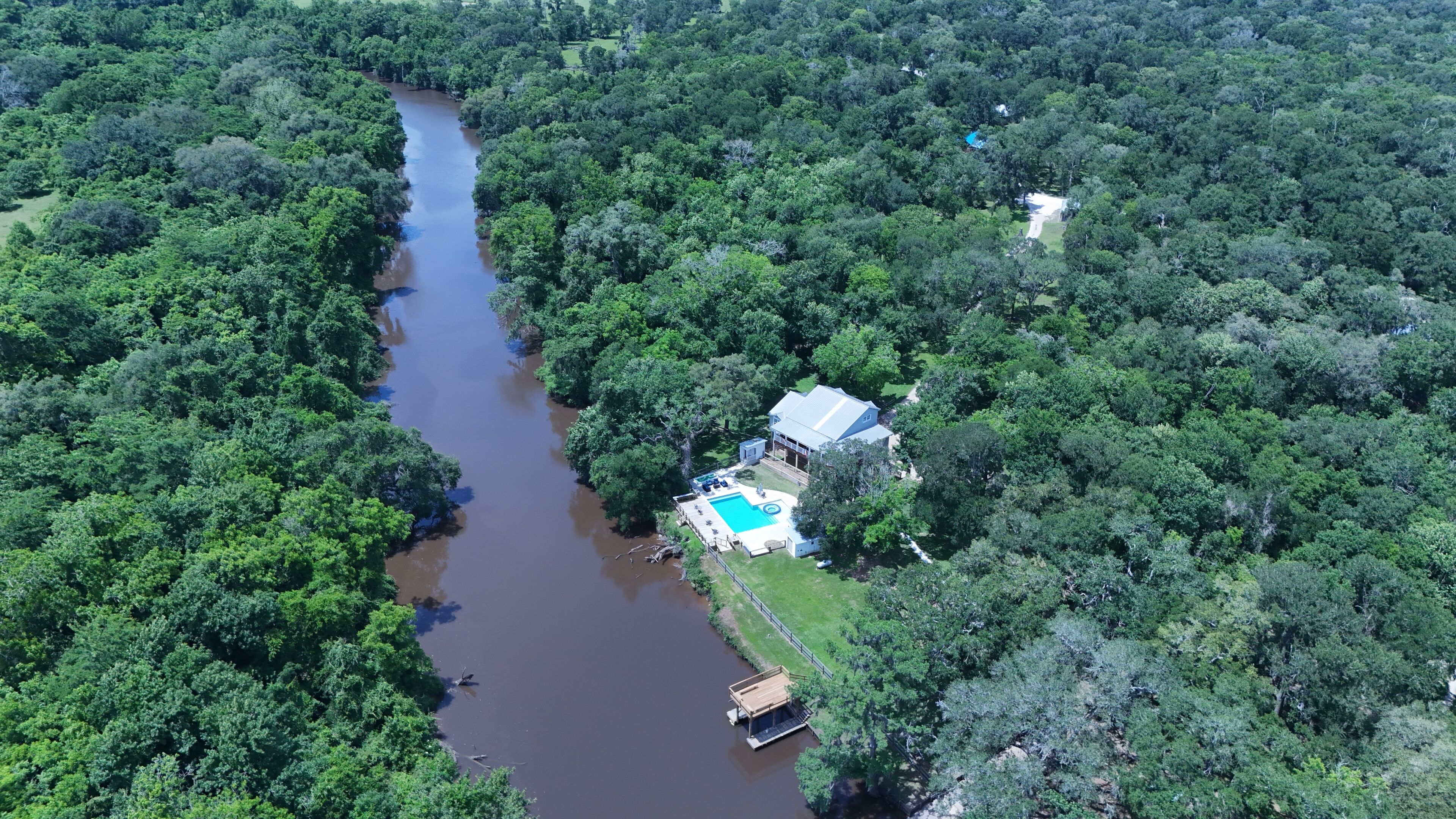 An aerial view of a riverside property with a swimming pool, dock, and lush greenery.