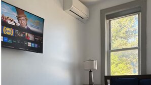 Quarto com cama, mesa de cabeceira, abajur, televisão fixada na parede e janela com vista para árvores.