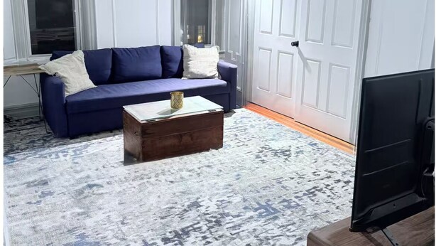 Sala de estar com sofá azul, mesa de centro de madeira, tapete estampado, televisão e porta branca.