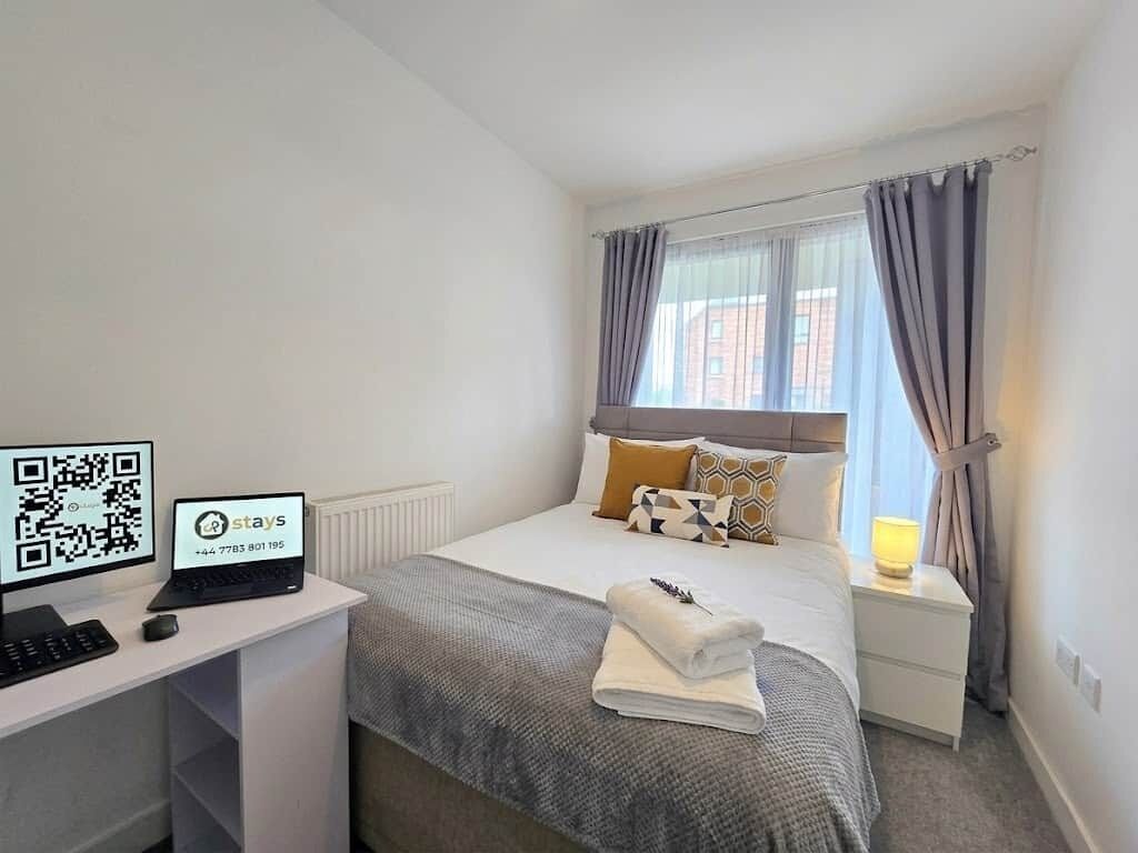 Une chambre d’hôtel comprenant un lit, un bureau avec un ordinateur portable et un écran, une lampe de chevet et une fenêtre avec des rideaux.