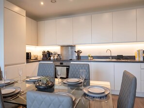 A modern kitchen with white cabinets, a stainless steel range hood, and a dining area with a glass table set for two. - Stylish 3Bed 2Bath: Quick Tubes to City & Wembley (London)