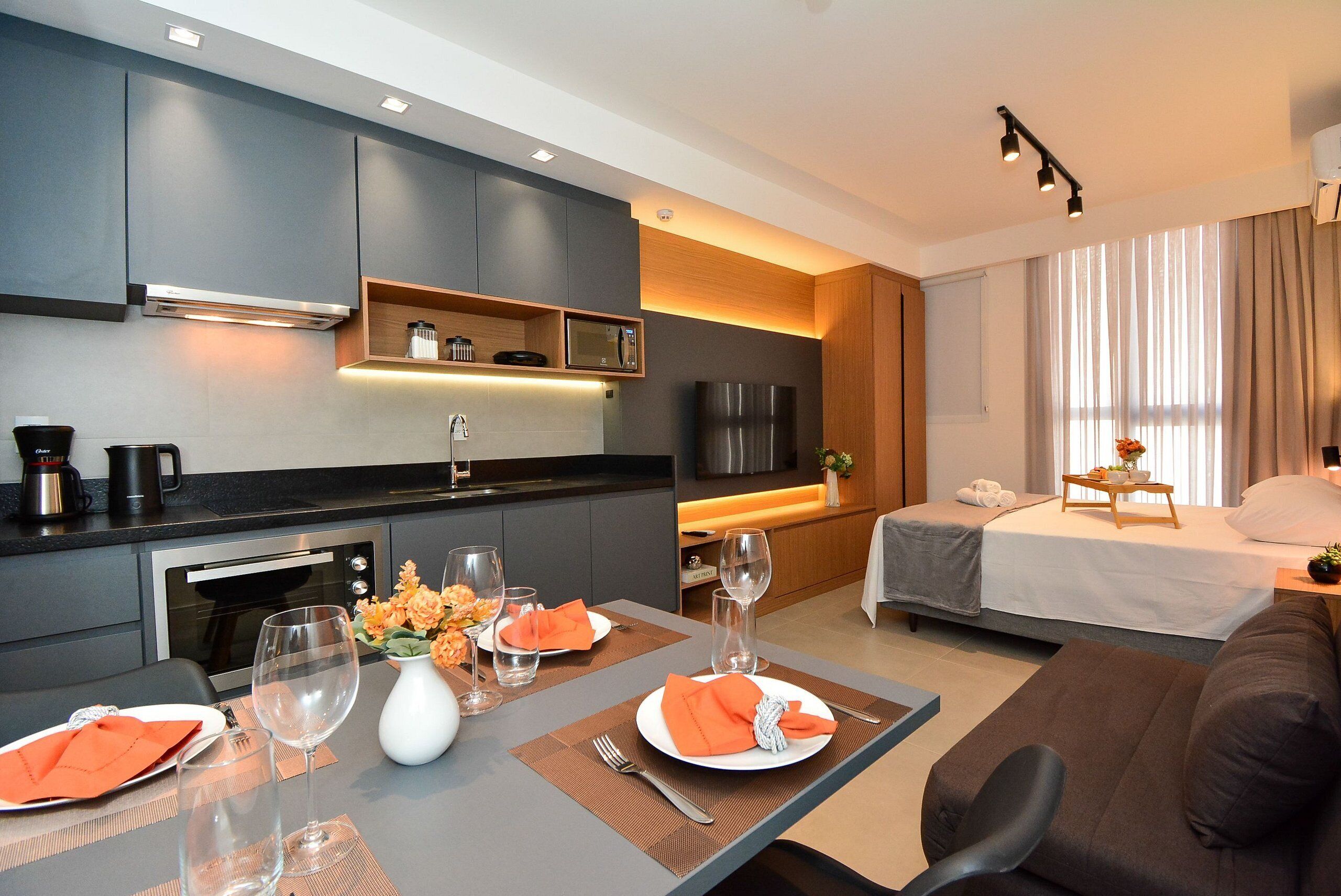 A modern hotel room with a kitchenette, a dining area, a bed, and a TV.