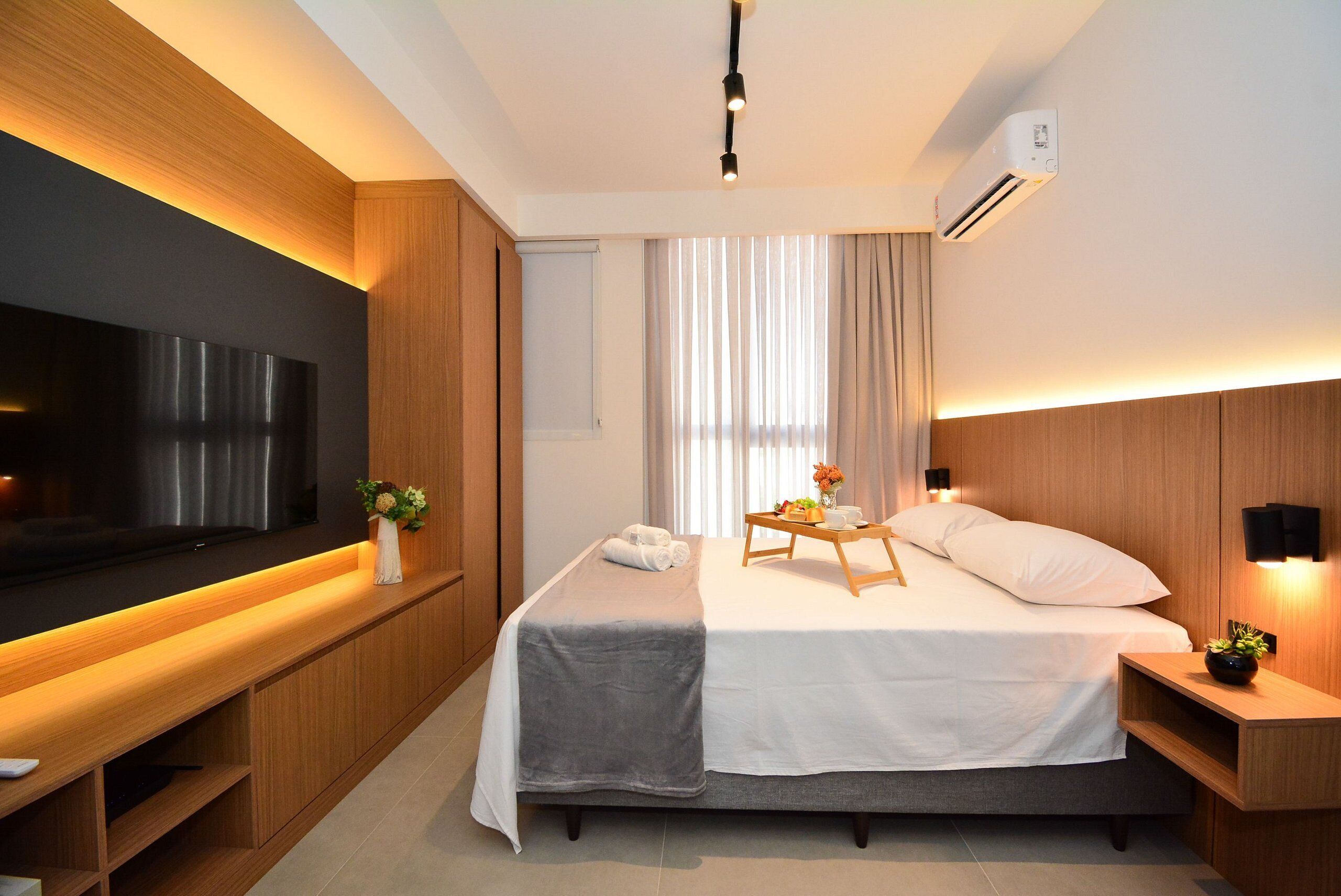 A modern hotel room with a large flat-screen TV, a bed with a bedside table, a wooden headboard, and a small desk area.