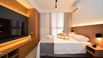 A modern hotel room with a large flat-screen TV, a bed with a bedside table, a wooden headboard, and a small desk area.
