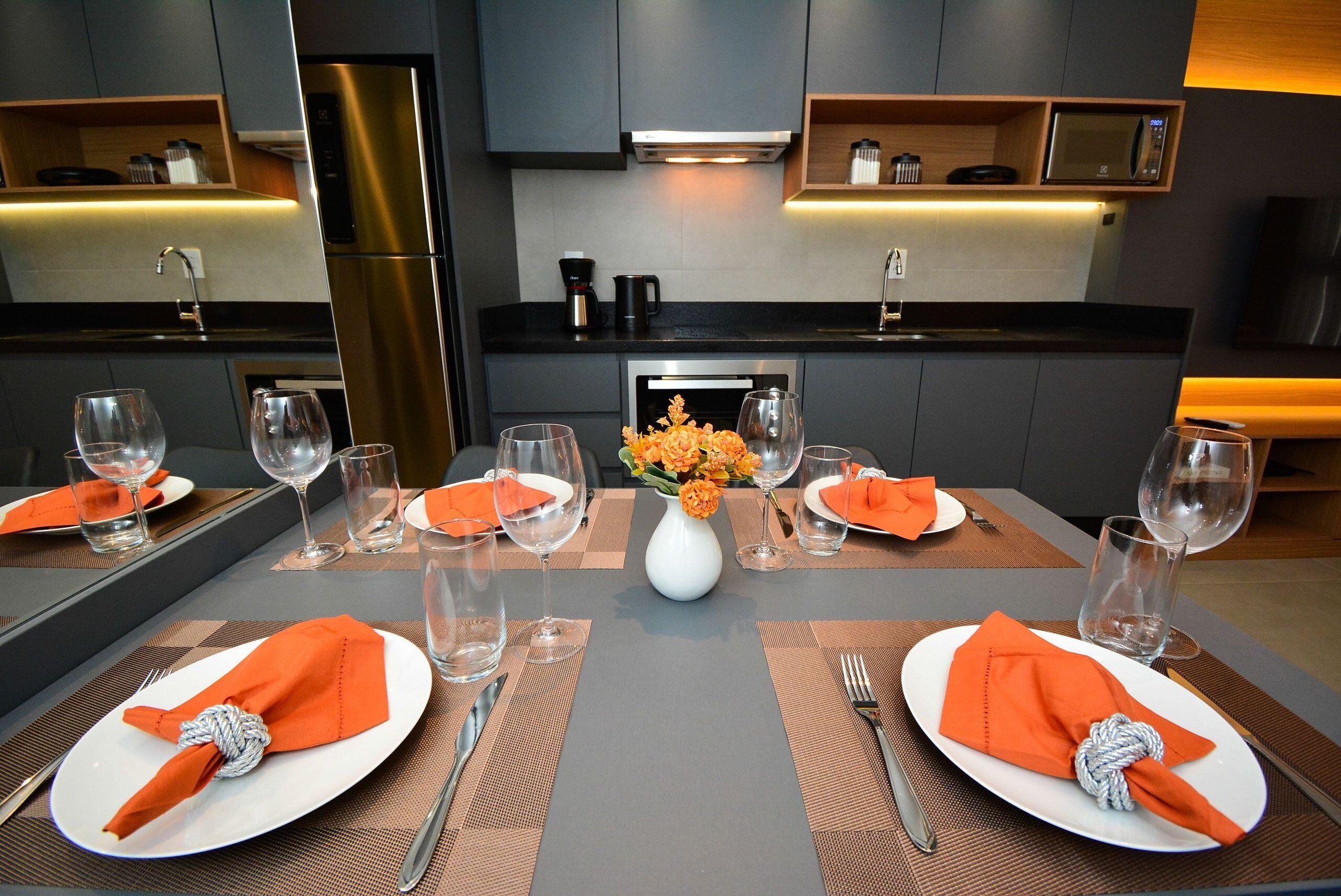 A modern dining area with a central table set for four, featuring a white vase with orange flowers, and a kitchen with a built-in microwave and stainless steel appliances in the background.