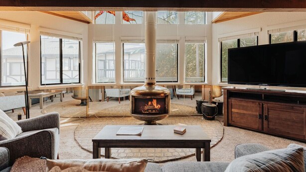 A cozy living room with a wood-burning stove, comfortable seating, and large windows. - Golden Hour | Jacuzzi, Sauna, Pool, Tennis, Gated! (Big Bear Lake)