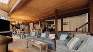 A spacious living area with a high ceiling, wooden beams, a television, a dining table, and a staircase. - Golden Hour | Jacuzzi, Sauna, Pool, Tennis, Gated! (Big Bear Lake)
