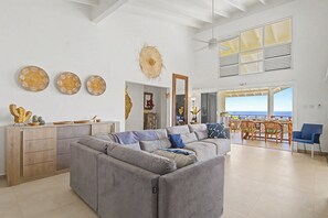 A spacious living area with a large gray sectional sofa, a wooden cabinet, and a dining area with a view of the sea.
