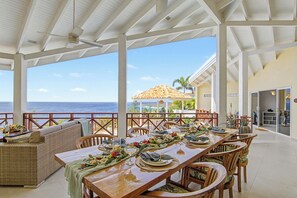 A covered dining area with a view of the ocean, a dining table set with plates, glasses, and a woven sofa.