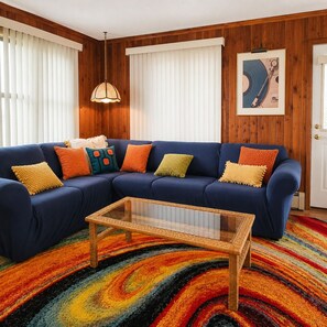 A blue sofa with colorful cushions, a glass coffee table, and a multicolored striped rug in a room with wooden paneling and white blinds. - Beach Front. The Plum Pad. Sunset, Surf & 70s Soul (Milton)