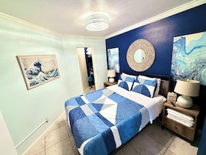 A bedroom with a bed, nightstands, a mirror, artwork, and a door leading to another room.
