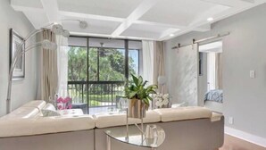 Un salon moderne avec un canapé, une table basse en verre et une vue sur la verdure à l’extérieur.
