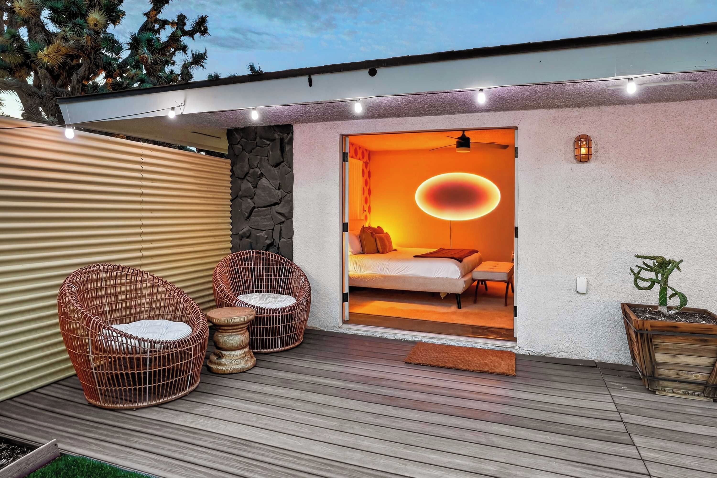 A modern outdoor space with a wooden deck, wicker chairs, and a cozy interior with a bed and ambient lighting.